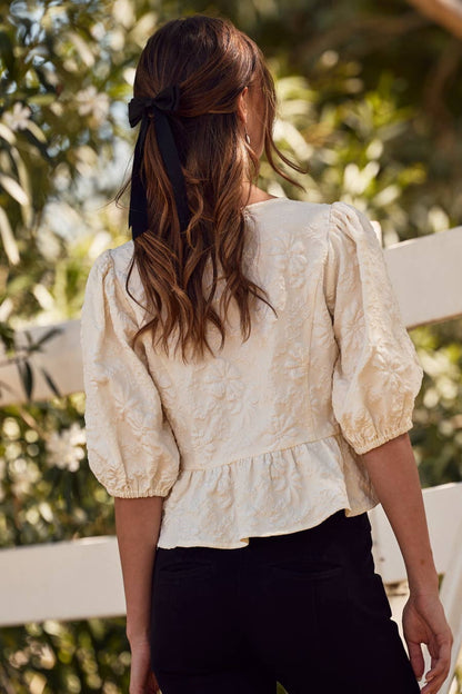 Woman wearing a beige lace blouse with black pants, standing outdoors with greenery in the background.