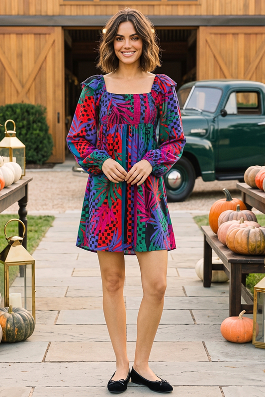 A woman stands outdoors in a rustic yard wearing a colorful, short puff-sleeve dress with a vibrant abstract pattern in pink, purple, blue, and green. She is wearing black flat shoes, with pumpkins and a green vintage pickup truck in the background.