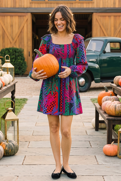 A woman stands outdoors in a fall-themed setting, holding a large orange pumpkin. She is wearing a colorful, long-sleeve mini dress with a geometric pattern in shades of purple, pink, green, and blue, paired with black flats.